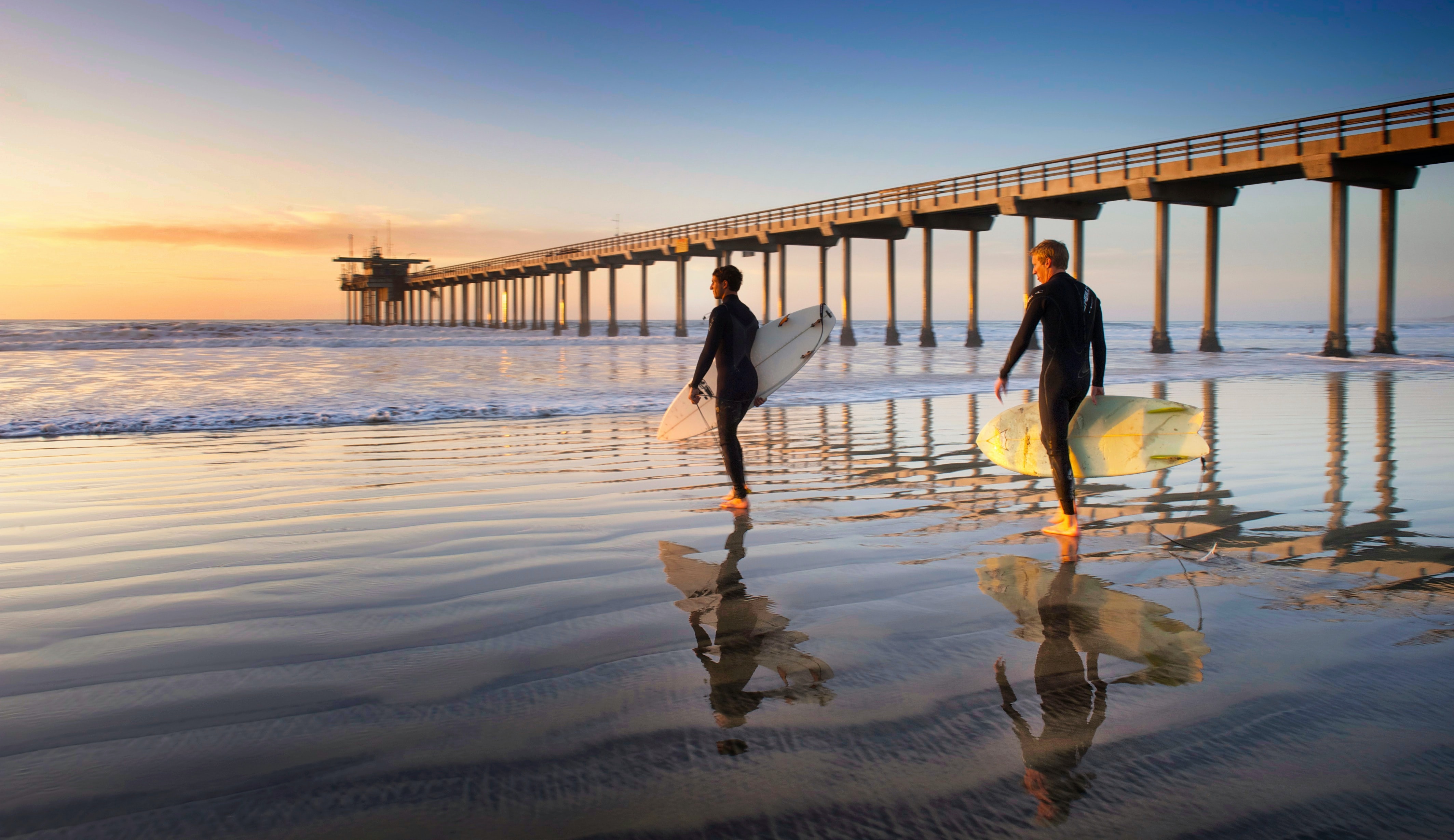 Two surfers walk along a reflective beach at sunset, carrying their surfboards near a long pier extending into calm waves. Ends with a period.