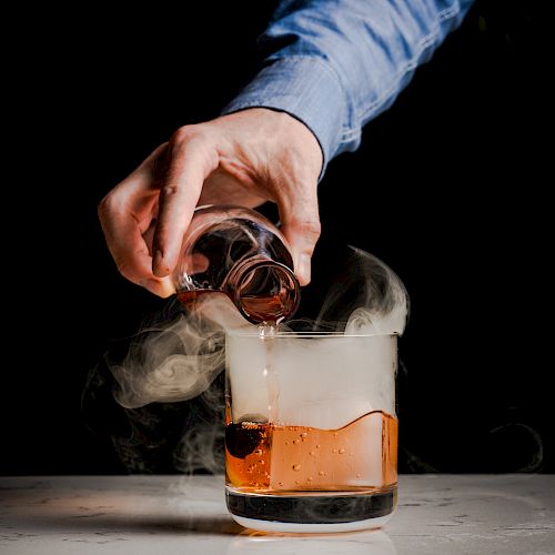 A hand pouring a dark liquid from a small glass into a larger glass emitting smoke, creating a visually intriguing effect.