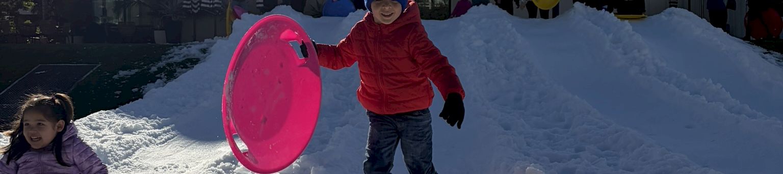 Kids play in a snowy courtyard, rolling on sledding tubes, with a modern building and sunny sky in the background.