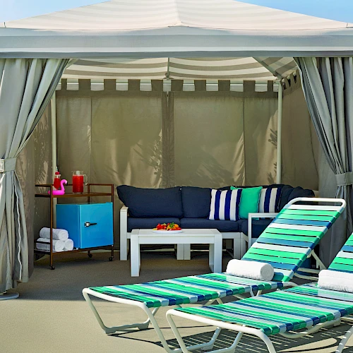 A poolside cabana with striped lounge chairs, a small table with a pink flamingo float, and a shaded area with a sofa and storage cabinet.