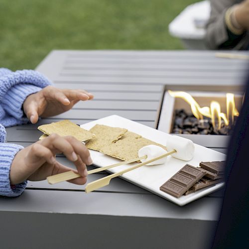 A person in a blue sweater holds skewers with marshmallows near a fire pit, preparing to make s'mores with graham crackers and chocolate on a plate.