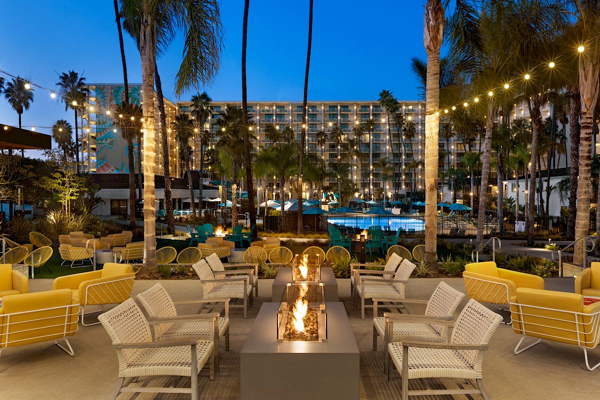 Resort courtyard at dusk with palm trees, lit string lights, teal umbrellas, and cozy fire pits before a hotel building.