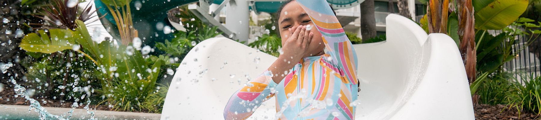 A child rides a water slide, shielding their face as the splash and water spray burst everywhere.