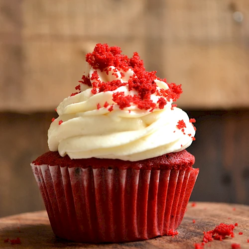 A red velvet cupcake with cream cheese frosting and red crumbs on top, sitting on a wooden surface.