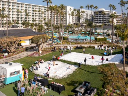 A sunny resort scene with palm trees and a pool, people setting up a large white base on the grass, tents and tables nearby.