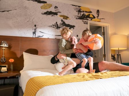 Three kids play on a hotel bed as a parent relaxes; beach mural, warm lamp glow, and yellow accents set a cozy scene.