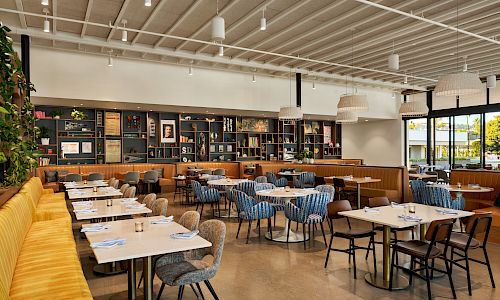 A bright, modern restaurant dining area with white tables, mixed blue and gray chairs, a long yellow sofa, and a back wall of shelves and windows.