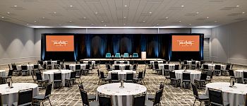 A large conference room set with round tables and chairs, a stage with orange screens, and ample lighting for a formal event.