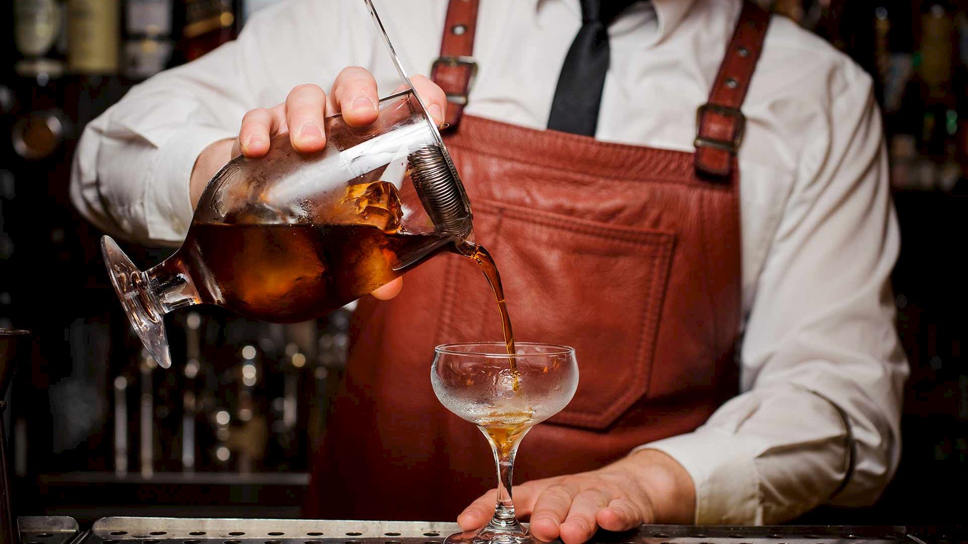 A bartender in a white shirt, black tie, and leather apron pours a cocktail from a mixing glass into a coupe glass.