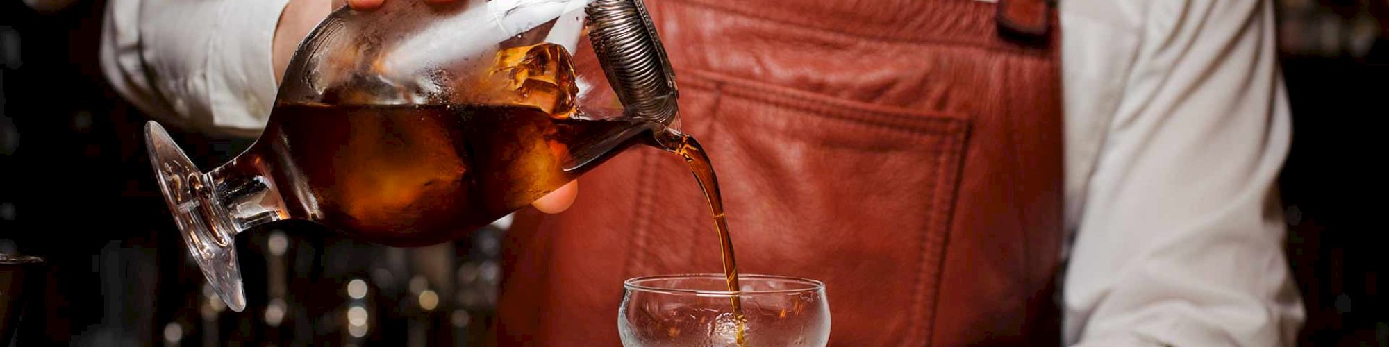 A bartender in a white shirt, black tie, and leather apron pours a cocktail from a mixing glass into a coupe glass.