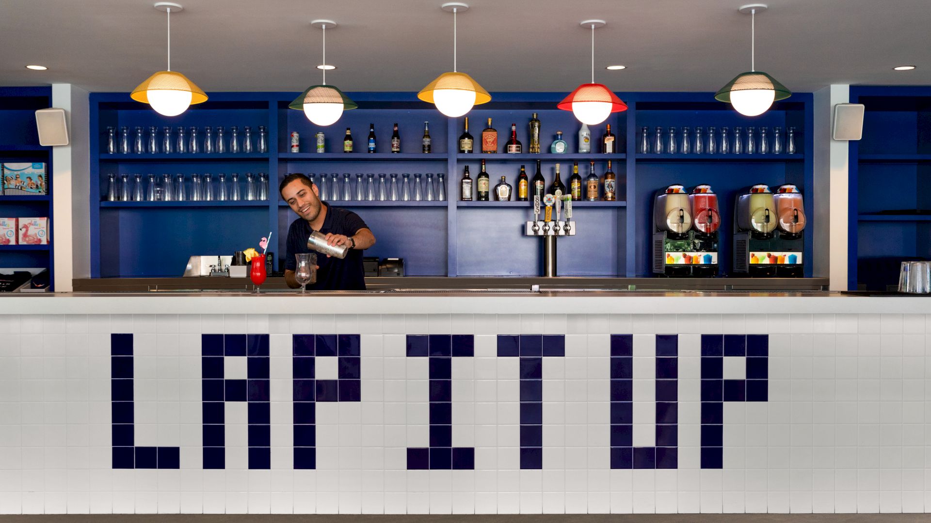 A bartender is working behind a bar with "LAP IT UP" spelled out in tiles on the front. There are bottles on shelves and drink machines.