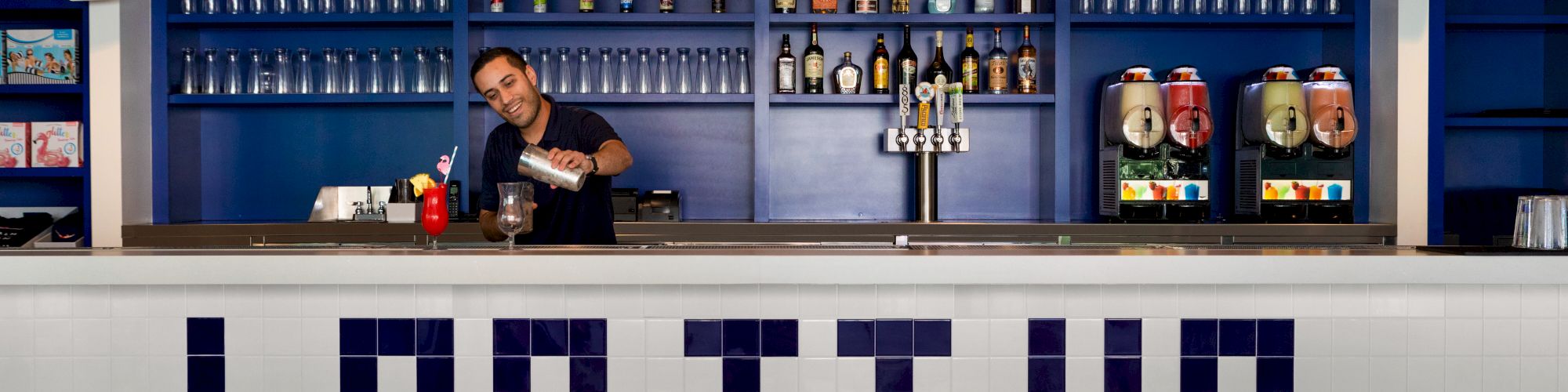 A bartender is working behind a bar with "LAP IT UP" spelled out in tiles on the front. There are bottles on shelves and drink machines.