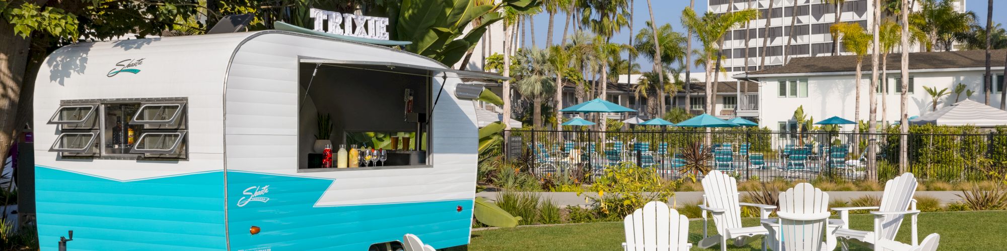 A retro-style camper is parked on a grassy area surrounded by white Adirondack chairs, with palm trees and a pool in the background.
