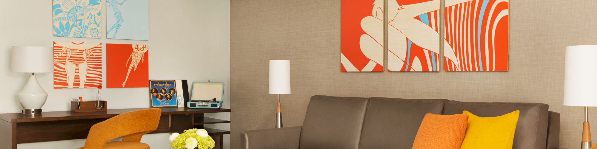 A modern living room with orange accents features a brown sofa, colorful artwork, a wooden coffee table, and a small desk.