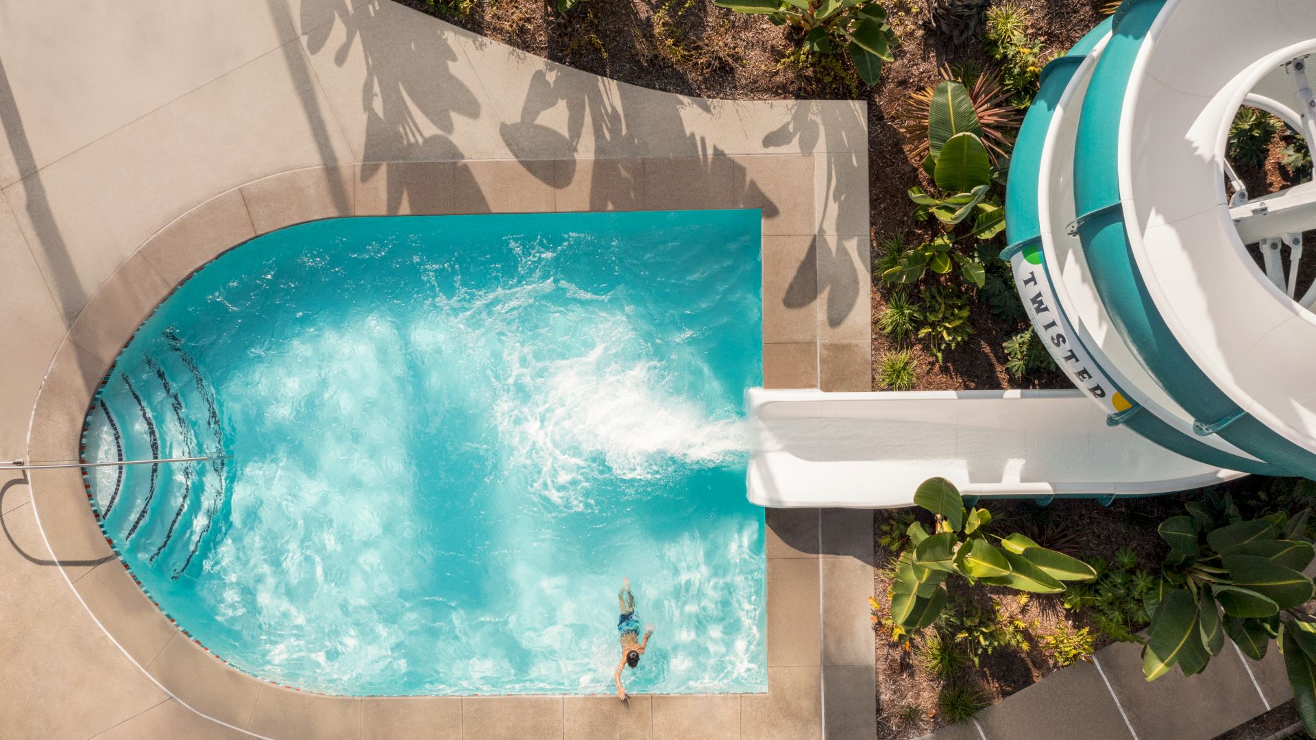The image shows a person at the end of a water slide, sliding into a small swimming pool surrounded by plants and paved walkways.