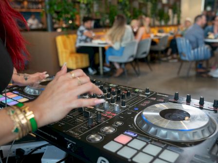 A DJ spins at a mixer and controllers, hands on the pad, clubgoers dining in the background, bright lights, lively mood.