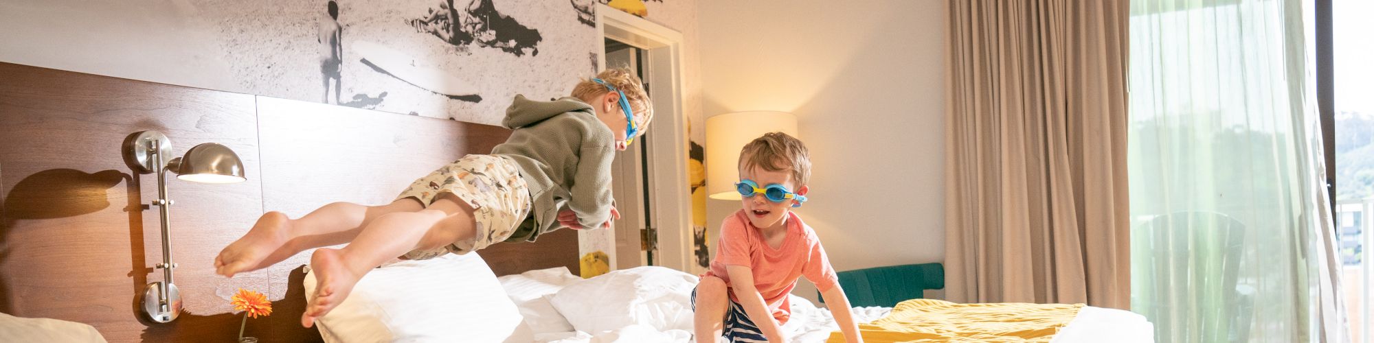 Two children wearing goggles are jumping on twin beds in a brightly lit hotel room.