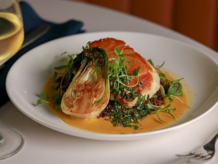 A fancy plated dish with a salmon fillet on greens, a fennel-like garnish, and a light orange sauce, plus a glass of white wine on the side.
