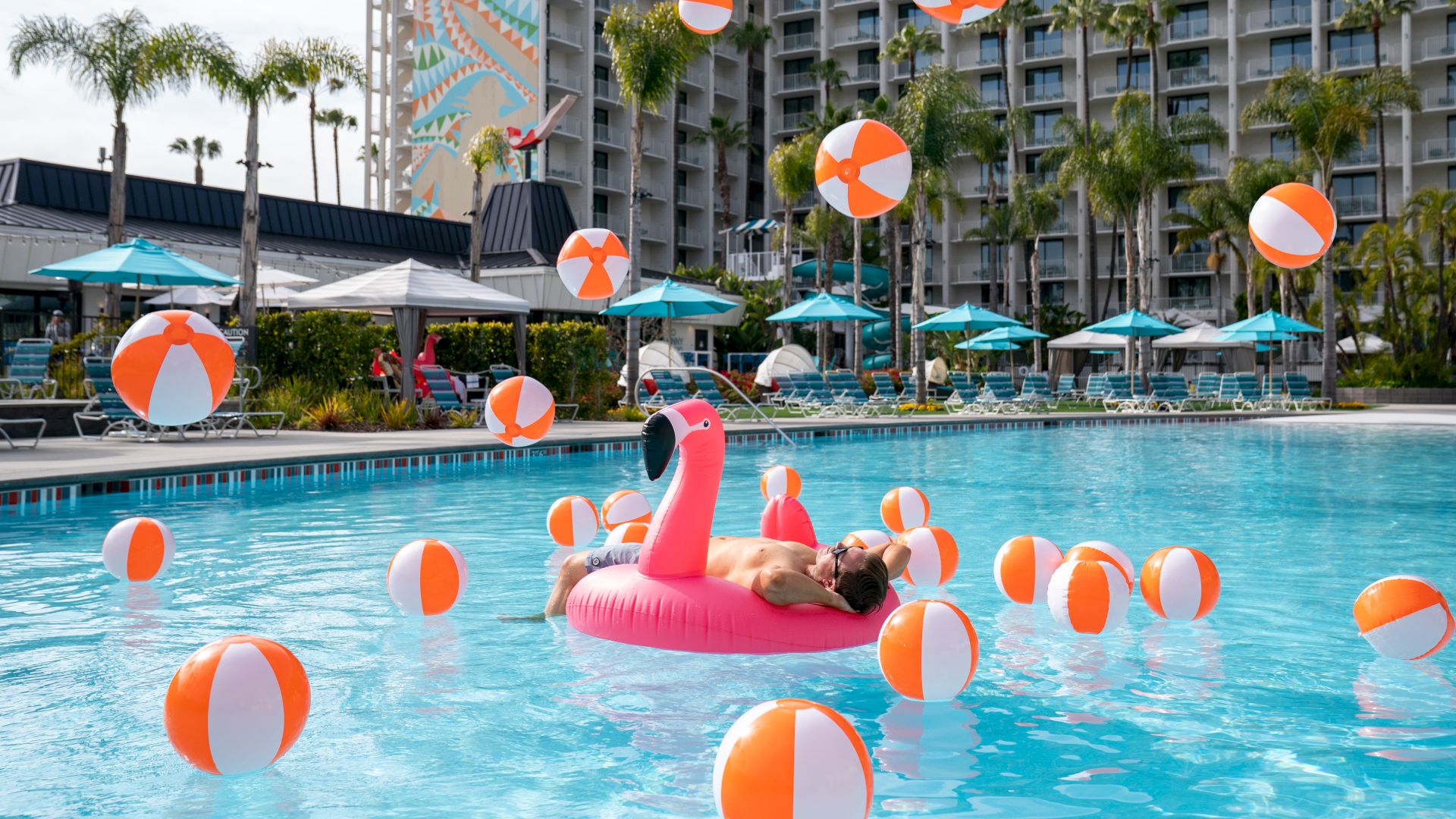 A man lounges in a pool with an inflatable flamingo and orange-white beach balls, set beside umbrellas and hotel rooms.