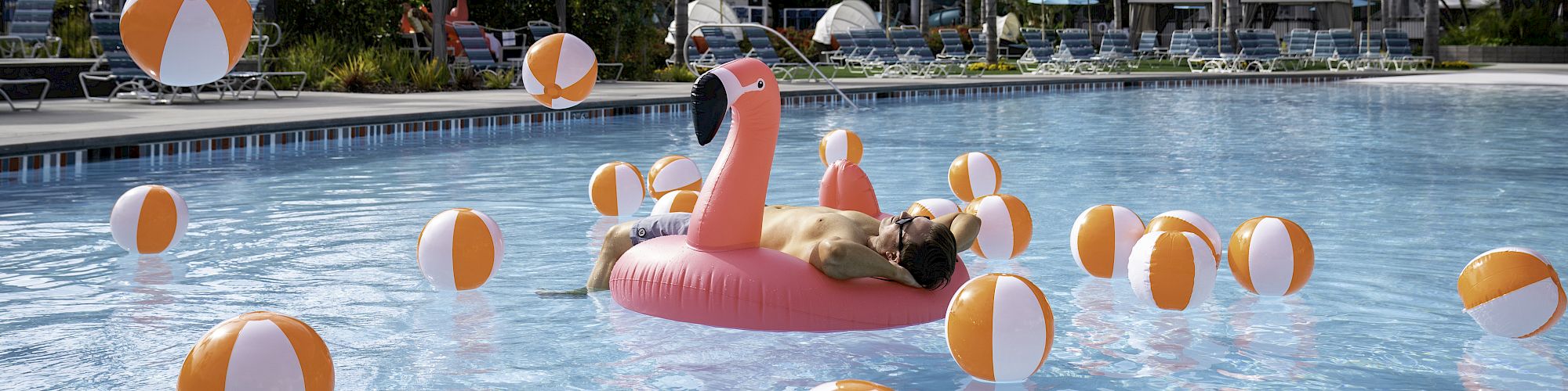 A swimming pool surrounded by a hotel with palm trees, a person on a pink flamingo float, and numerous yellow and white beach balls in the water.