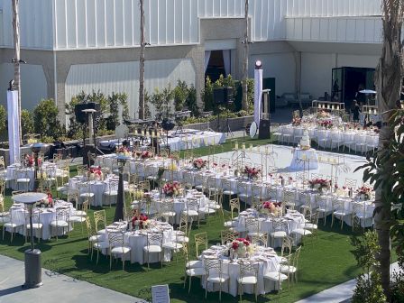 A formal outdoor event setup with many round tables, white chairs, floral centerpieces, and banquet arrangements on a lawn near a building.
