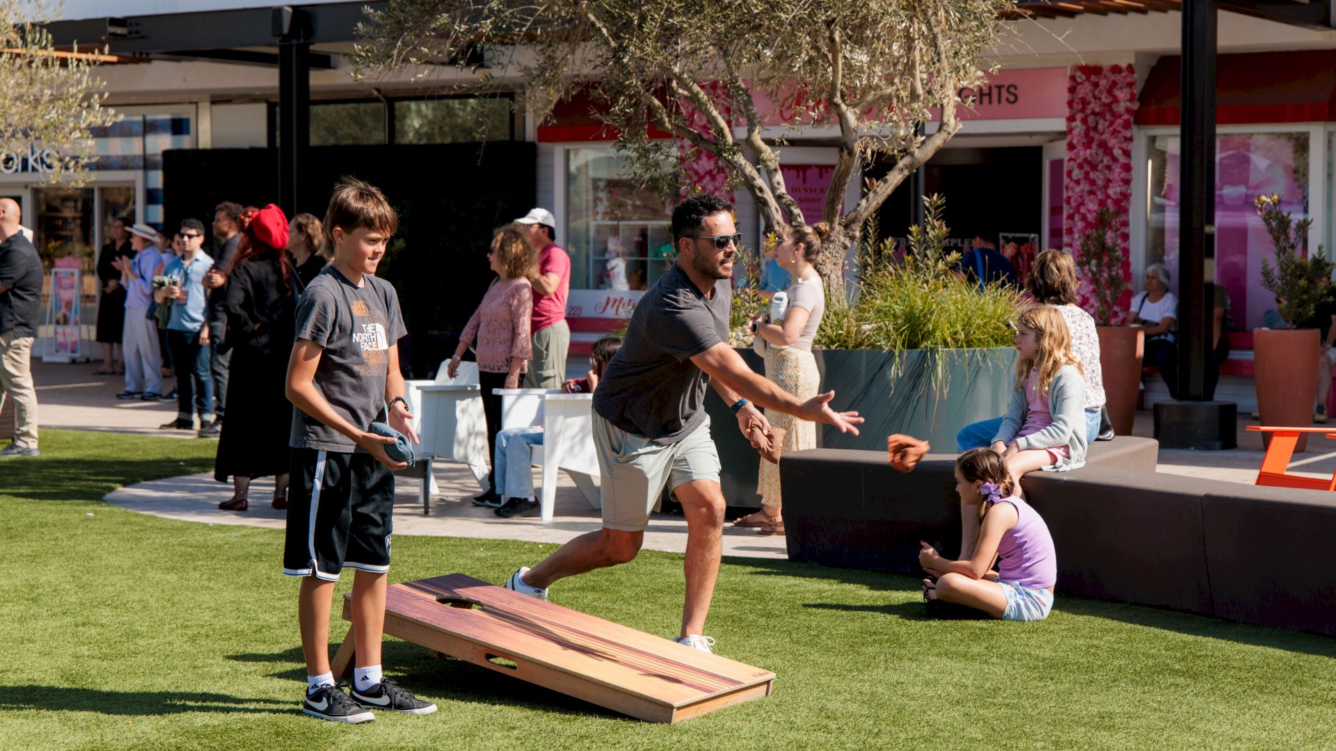 Kids and adults enjoy a sunny outdoor cornhole-style game on the lawn beside a shopping center.