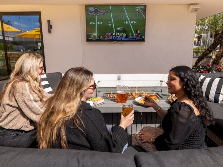 Three friends dine on a patio couch, watching a football game on a wall-mounted TV while sipping drinks and sharing snacks.