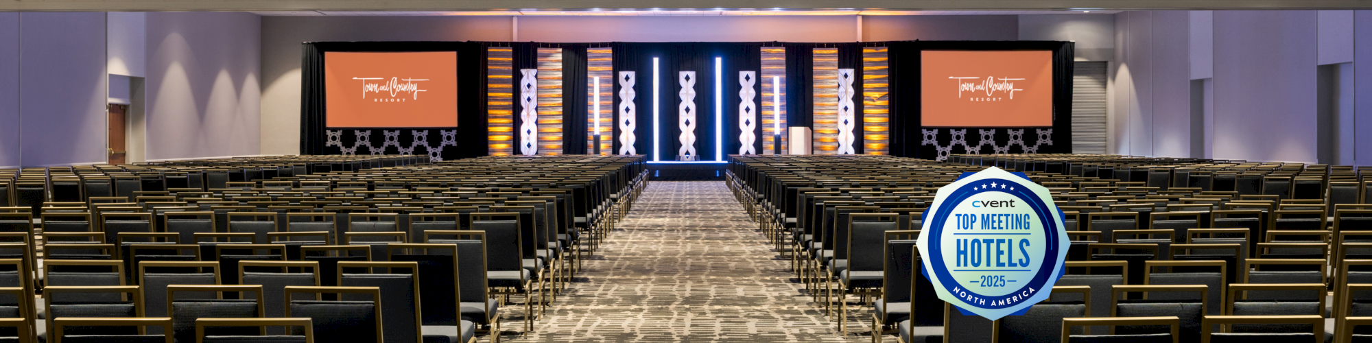 The image shows an empty conference room with rows of chairs and a stage at the front, featuring a "Top Meeting Hotels" badge.