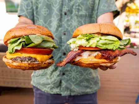 Two people hold two large burgers with lettuce, cheese, tomato, bacon and patties in a casual outdoor setting.