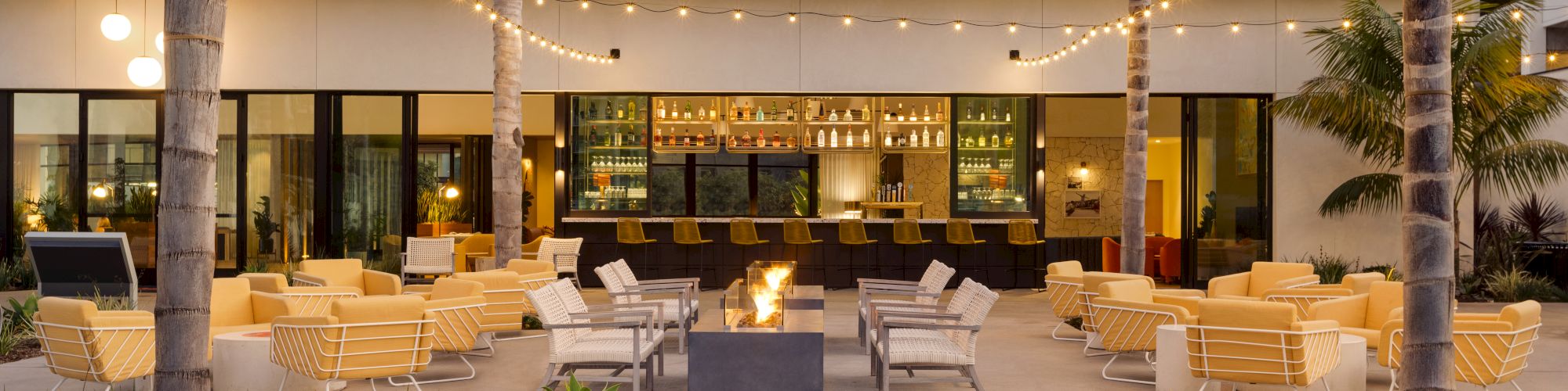 An outdoor lounge area features yellow seating, palm trees, string lights, and a central fire pit, adjacent to a well-lit bar with bottles on display.