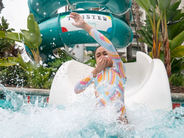 A person in a colorful wetsuit rides a splashy water slide into a pool, with a teal water slide twisty tube in the background.
