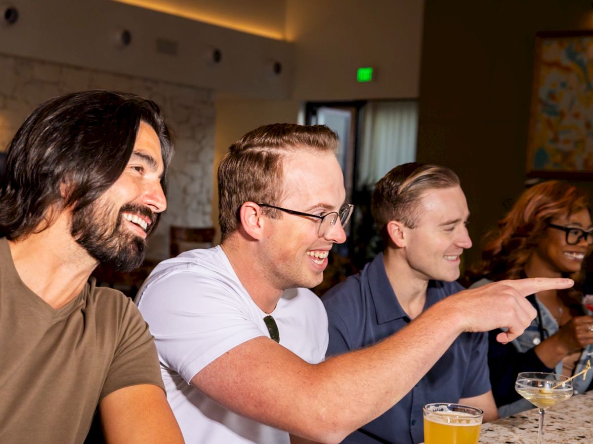 Three men sit at a bar counter, smiling and chatting. The man in white points as the others listen, with drinks in front of them and warm, modern lighting overhead.