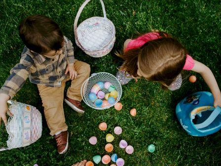 Two kids Easter egg hunting on the grass, baskets full of colorful eggs and a blue helmet nearby, sunny outdoor scene.