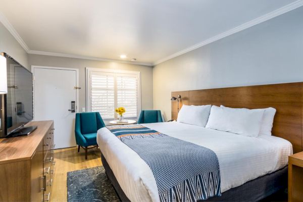 A modern hotel room features a king-sized bed with white linens, a wooden headboard, two blue chairs, a small table with flowers, and a flat-screen TV. Best resort in san diego area
