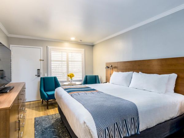 A modern hotel room features a king-sized bed with white linens, a wooden headboard, two blue chairs, a small table with flowers, and a flat-screen TV. Best resort in san diego area