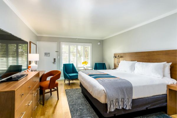 The image shows a modern hotel room with a large bed, wooden furniture, a desk, two blue chairs, a flat-screen TV, and large windows. San Diego hotels.
