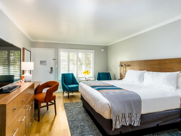 The image shows a modern hotel room with a large bed, wooden furniture, a desk, two blue chairs, a flat-screen TV, and large windows. San Diego hotels.
