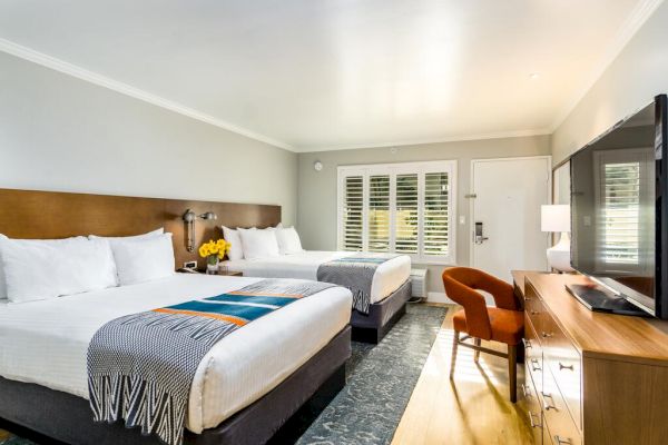 The image shows a well-lit hotel room with two double beds, a flat-screen TV, a desk, a chair, and a window with shutters. The beds have white linens. Hotels in San Diego