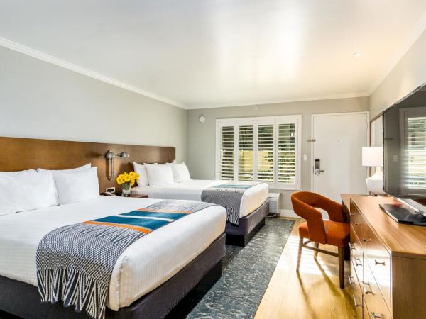 The image shows a well-lit hotel room with two double beds, a flat-screen TV, a desk, a chair, and a window with shutters. The beds have white linens. Hotels in San Diego
