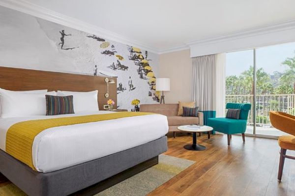 A hotel room with a king-size bed, modern decor, a wall mural, seating area, desk, large window, and a view of greenery outside. San Diego hotel