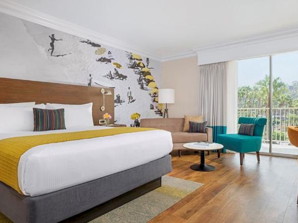 A hotel room with a king-size bed, modern decor, a wall mural, seating area, desk, large window, and a view of greenery outside. San Diego hotel