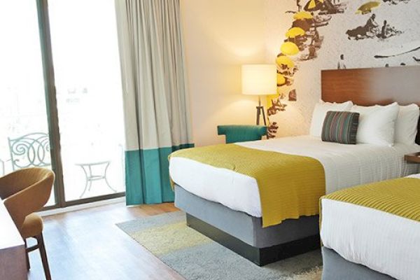 A hotel room with two beds, a decorative wall, a lamp, a chair, a desk, and a sliding glass door leading to a balcony with furniture. Hotels near San Diego Zoo