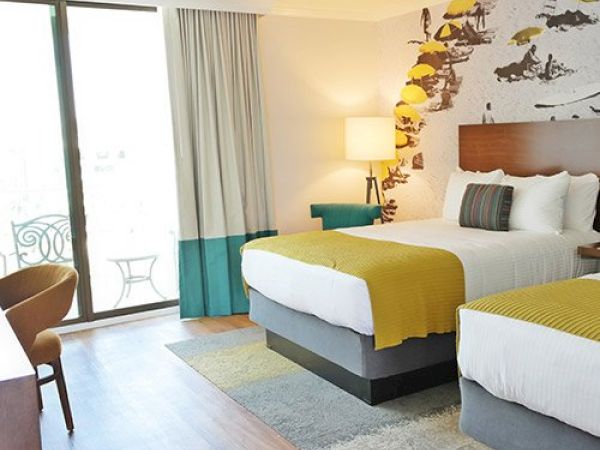 A hotel room with two beds, a decorative wall, a lamp, a chair, a desk, and a sliding glass door leading to a balcony with furniture. Hotels near San Diego Zoo