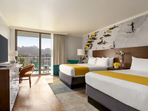 Two double beds in a bright hotel room with a mural of airplanes, a desk, a chair, a TV, and a balcony with a view.