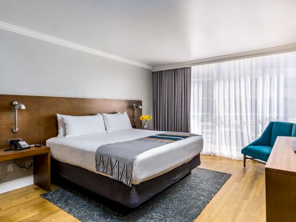A modern hotel room features a large bed, a TV, an armchair, and floor-to-ceiling windows with curtains, creating a bright and welcoming atmosphere. San Diego hotel