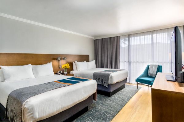 The image shows a hotel room with two neatly made double beds, a TV, an armchair, and a window with curtains.