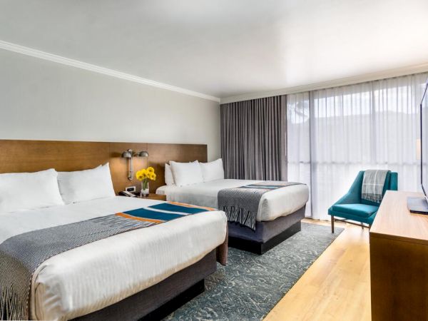 The image shows a hotel room with two neatly made double beds, a TV, an armchair, and a window with curtains.