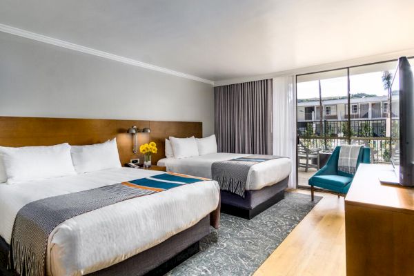 This image shows a hotel room with two double beds, a blue chair, a dresser, and a TV. There's a large window with a balcony view outside.