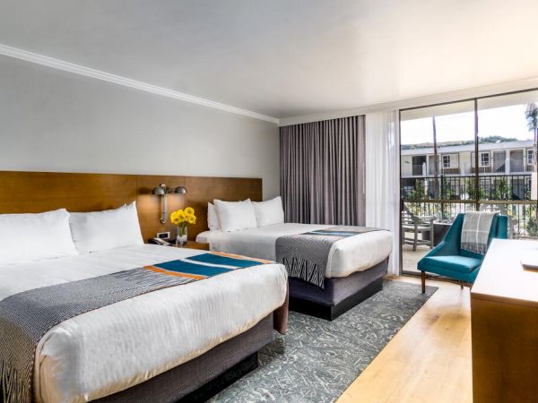 This image shows a hotel room with two double beds, a blue chair, a dresser, and a TV. There's a large window with a balcony view outside.