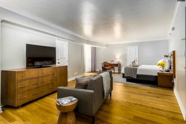 A spacious hotel room with a bed, TV on a dresser, sofa, small table, desk with chair, and wooden flooring. The room has soft lighting and minimal decor.
